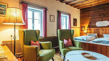 Ein Zimmer mit zwei grünen Sesseln, einem Holztisch, einem Bett mit karierter Bettwäsche, einem Heizkörper und einem Fenster mit Vorhängen.