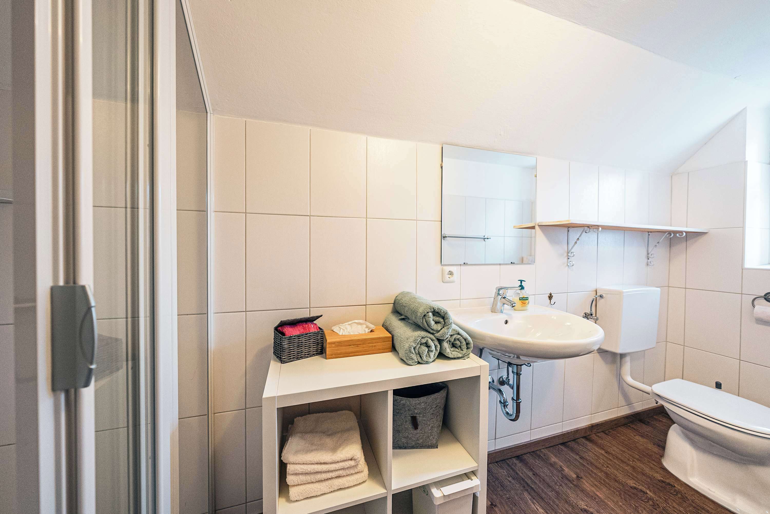 A modern bathroom with a white sink, toilet, and shower area. There is a shelf with towels and toiletries.