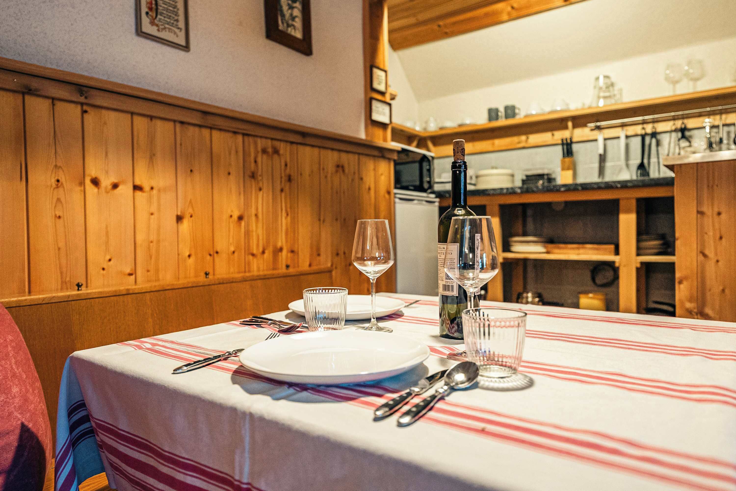 Ein Esstisch mit Tellern, Gläsern und Besteck, Holzvertäfelung an den Wänden und eine Küche im Hintergrund.