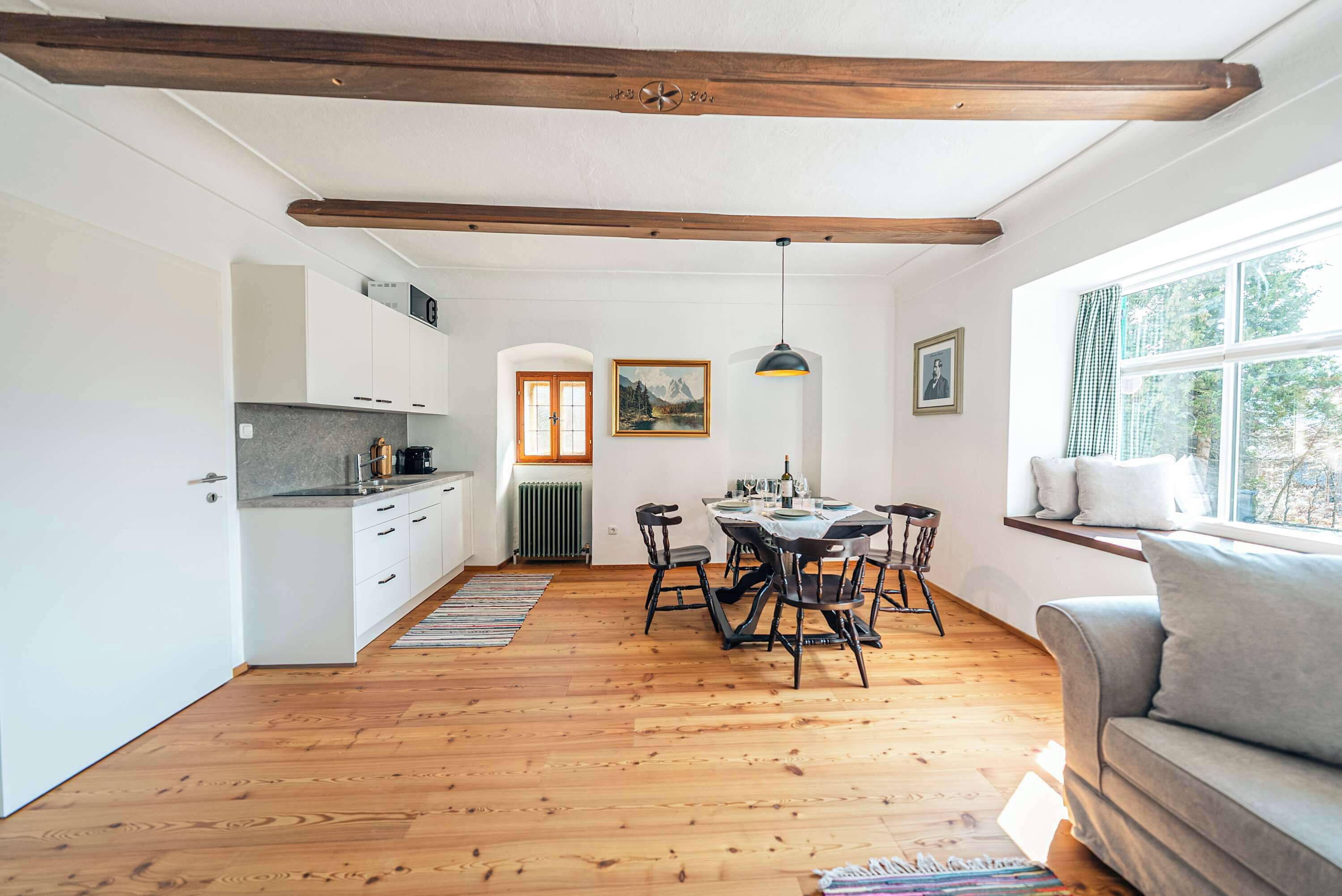 Cozinha moderna com armários brancos, área de jantar com mesa e cadeiras, vigas de madeira no teto e janela com vista para árvores.