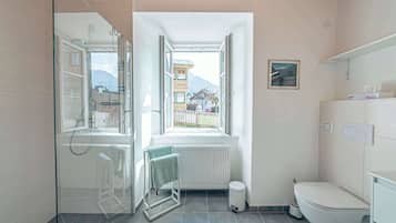 Une salle de bain moderne dotée d’une douche à parois de verre, de toilettes et d’une fenêtre offrant une vue sur les bâtiments et les montagnes.