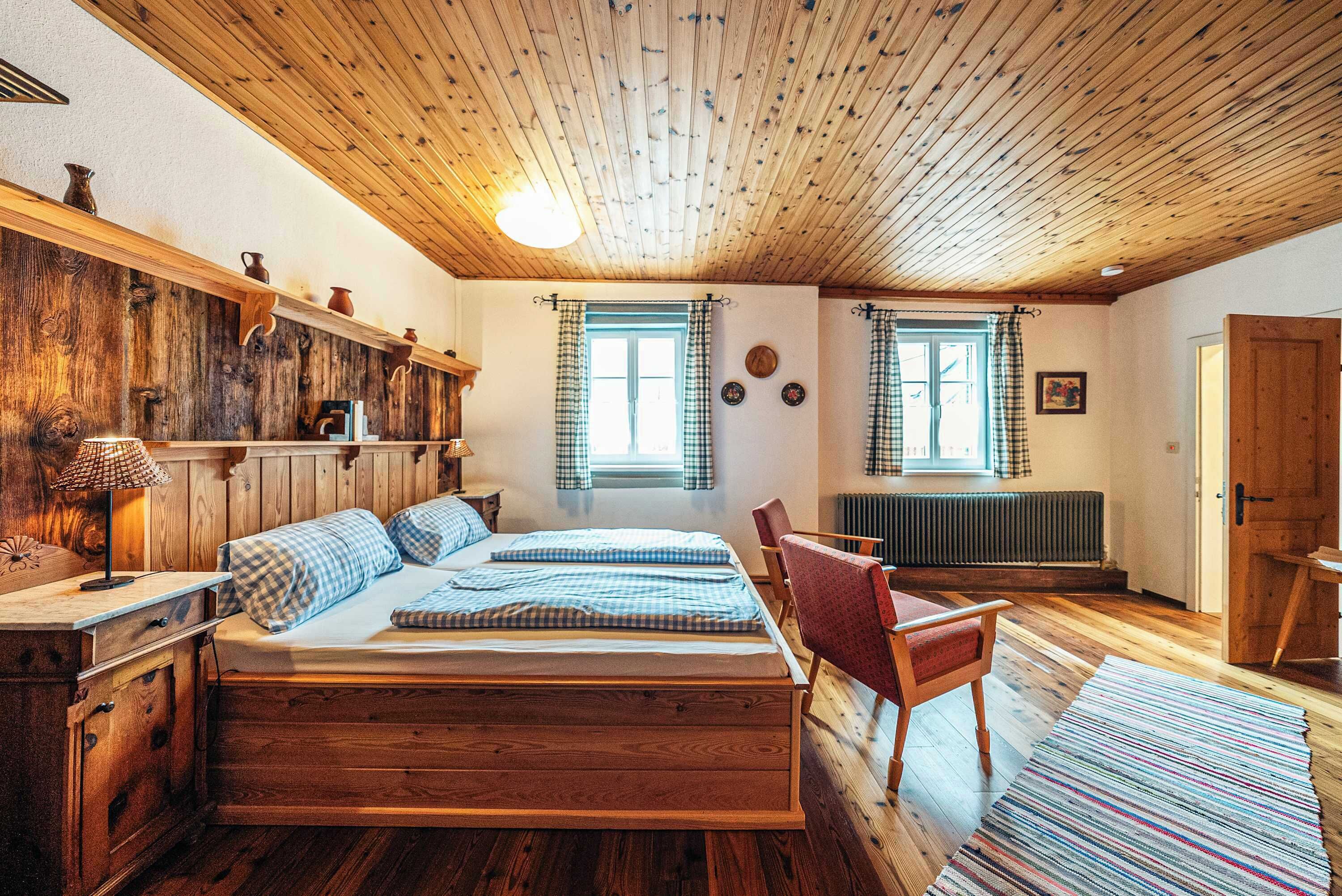 Ein Schlafzimmer aus Holz mit einem Bett, einem Sessel, einer Kommode und zwei Fenstern mit Vorhängen.