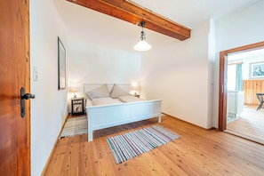 Un dormitorio con cama, mesita de noche, lámpara, suelo de madera, techo con vigas a la vista y una puerta.