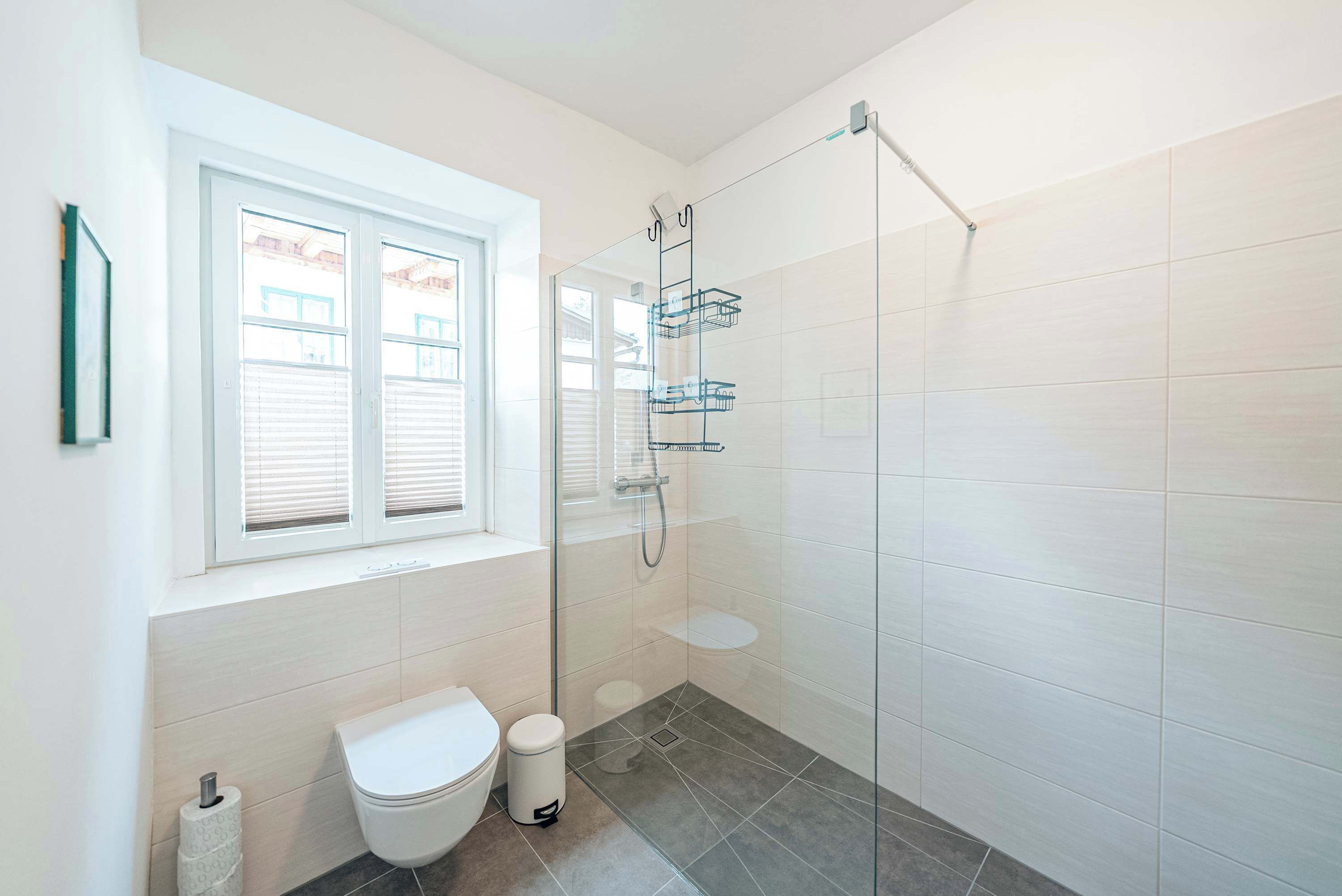 A modern bathroom with a glass shower enclosure, a toilet, and a window with blinds.
