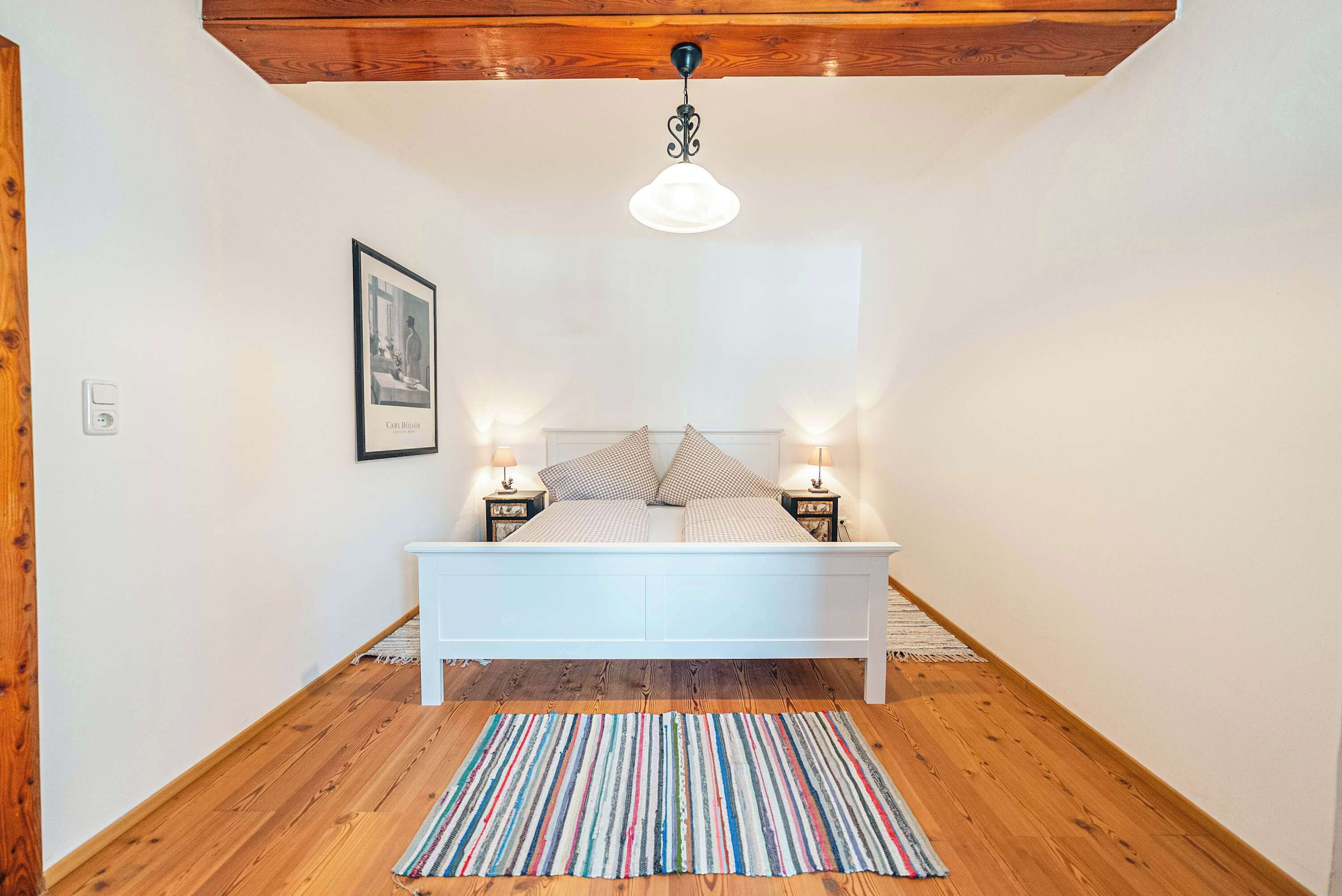 Ein Schlafzimmer mit einem weißen Bett, einem Streifen Teppich, einer Holzbalkendecke und zwei Nachttischlampen.