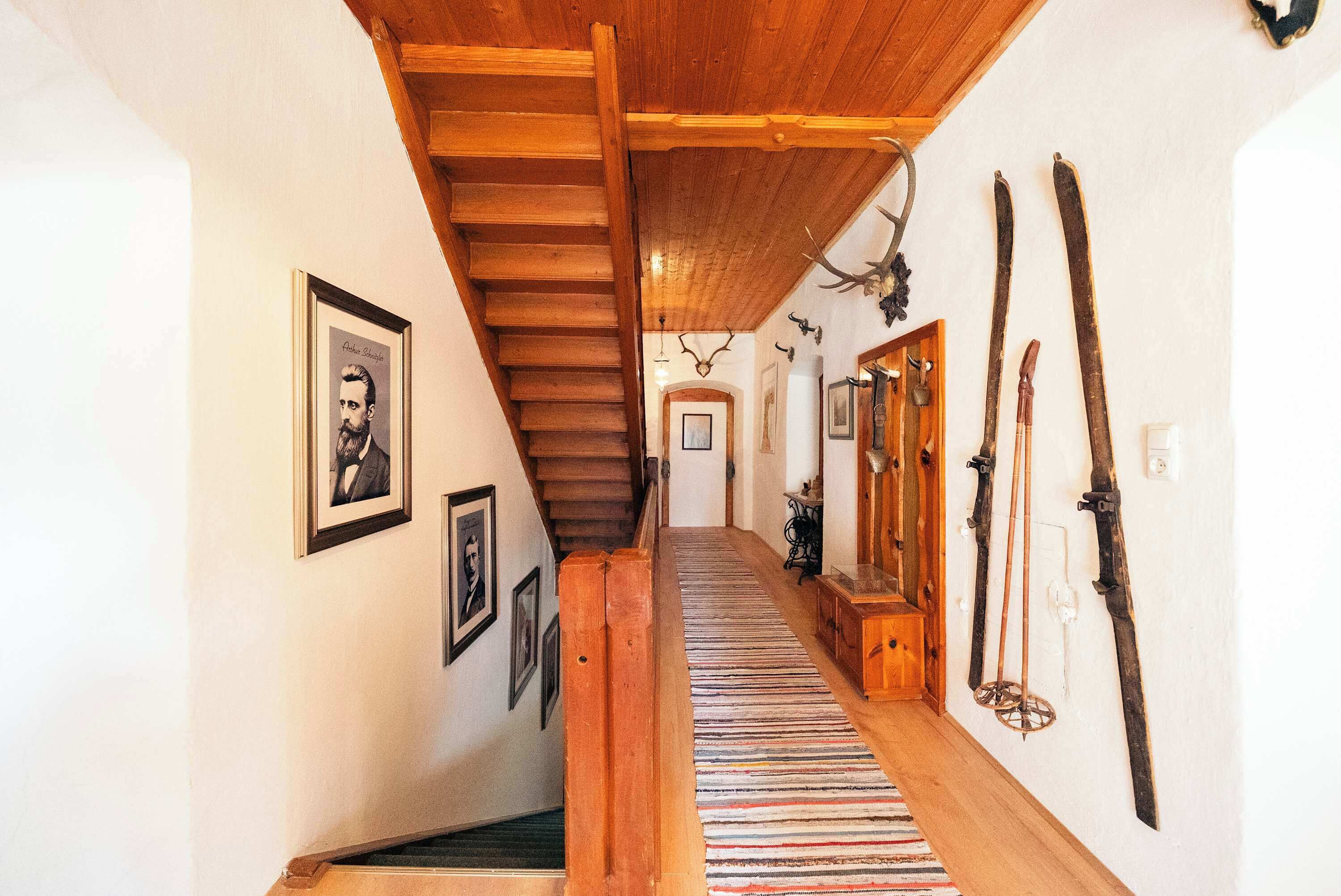 Eine Holztreppe führt zu einem Raum mit einem gerahmten Porträt, einem Streifen Teppich und an der Wand befestigten Ski.