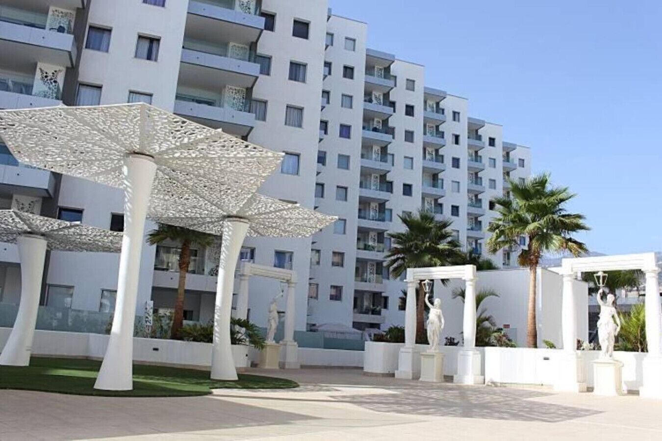 Un edificio moderno con balcones, sombrillas blancas y estatuas en un patio.