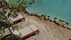 Deux fauteuils de jardin en bois, dotés de coussins beige, sont placés sur un sol de gravier, offrant une vue sur une étendue d’eau turquoise. Un mur de pierre et de la végétation se trouvent au premier plan.