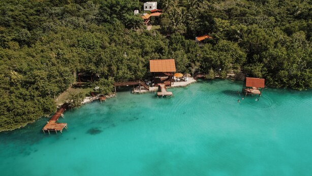 Vue aérienne d’une propriété située sur la côte, avec un quai en bois, un petit pavillon et une vaste zone ouverte entourée d’une végétation luxuriante.