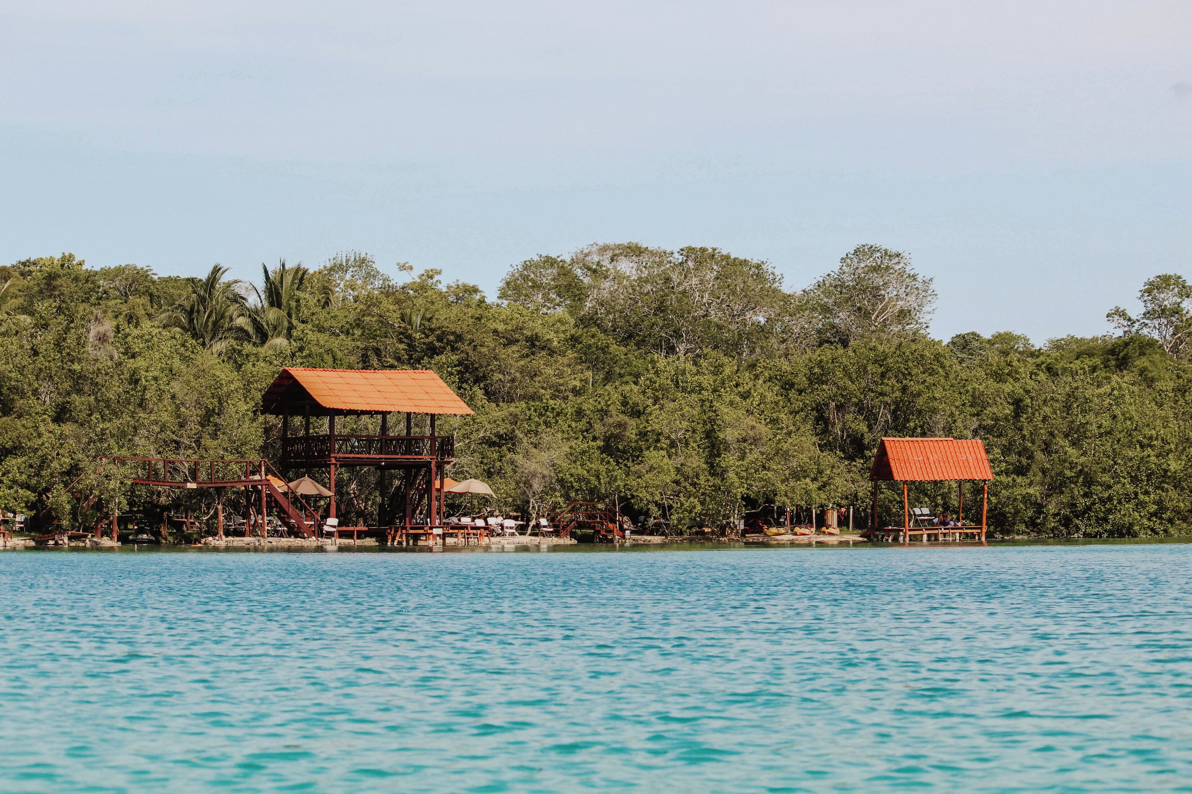 A wooden structure with a red roof by a clear blue lake, surrounded by dense green foliage.