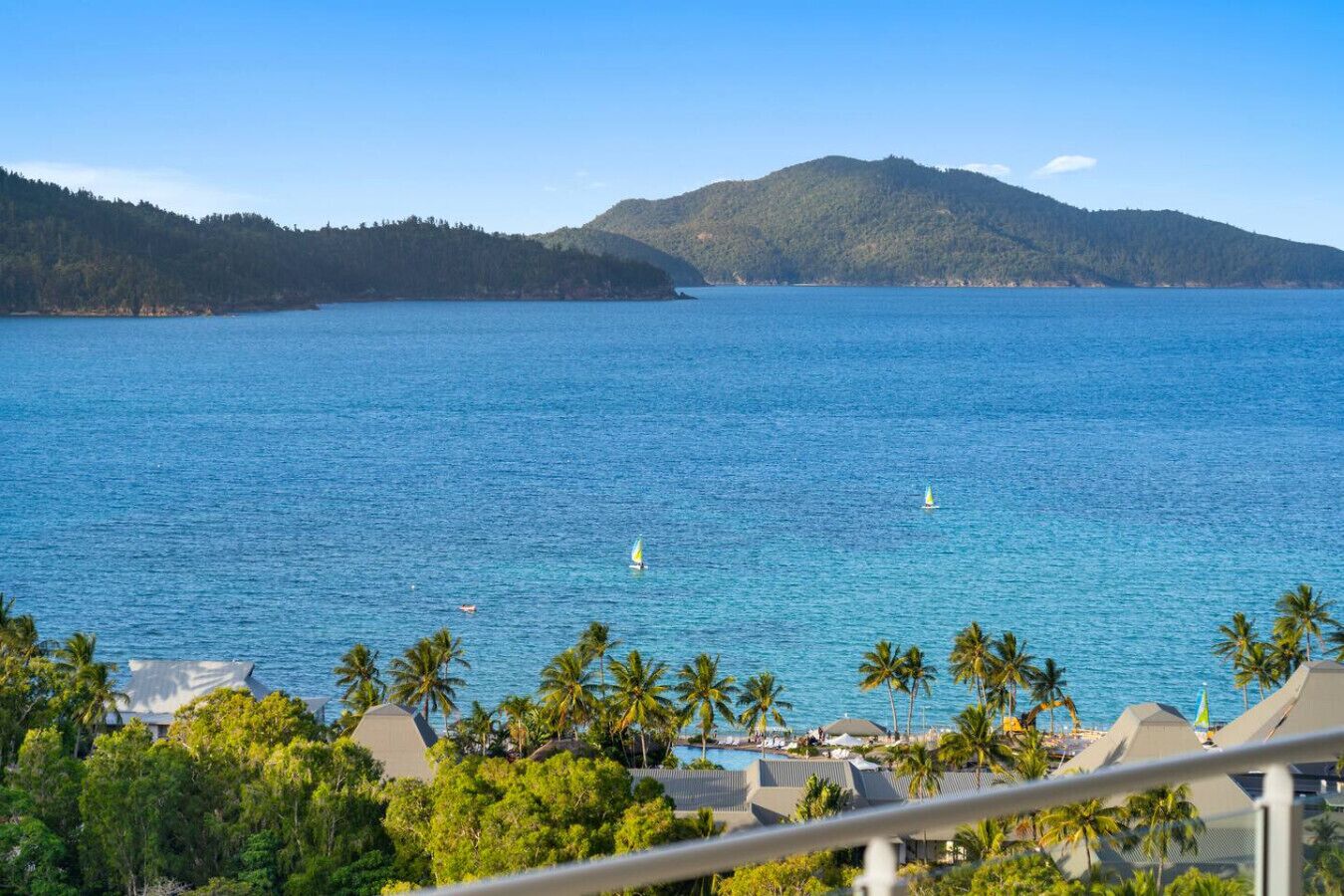 A coastal view with a clear blue sea, palm trees, and a distant mountain range.