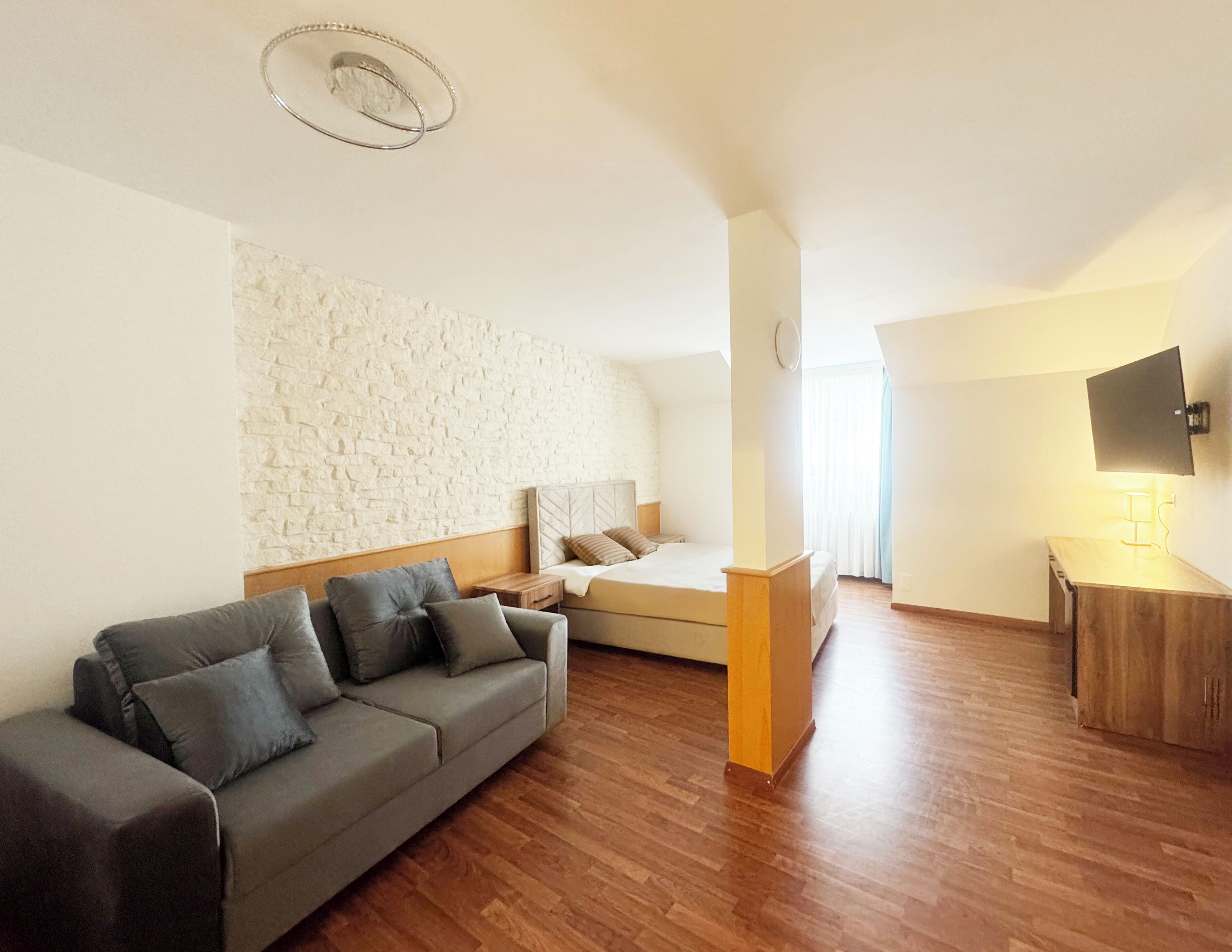 Una habitación de hotel moderna con una pared de acento de piedra, suelo de madera, sofá gris, cama con cabecera, mesita de noche, televisor montado en la pared y un pequeño mueble de madera.