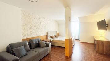 Una habitación de hotel moderna con una pared de acento de piedra, suelo de madera, sofá gris, cama con cabecera, mesita de noche, televisor montado en la pared y un pequeño mueble de madera.