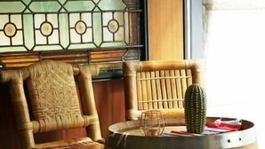 Un taburete de barril de madera con un cactus encima, colocado frente a una silla con respaldo de mimbre, cerca de una ventana con un diseño de vitral.