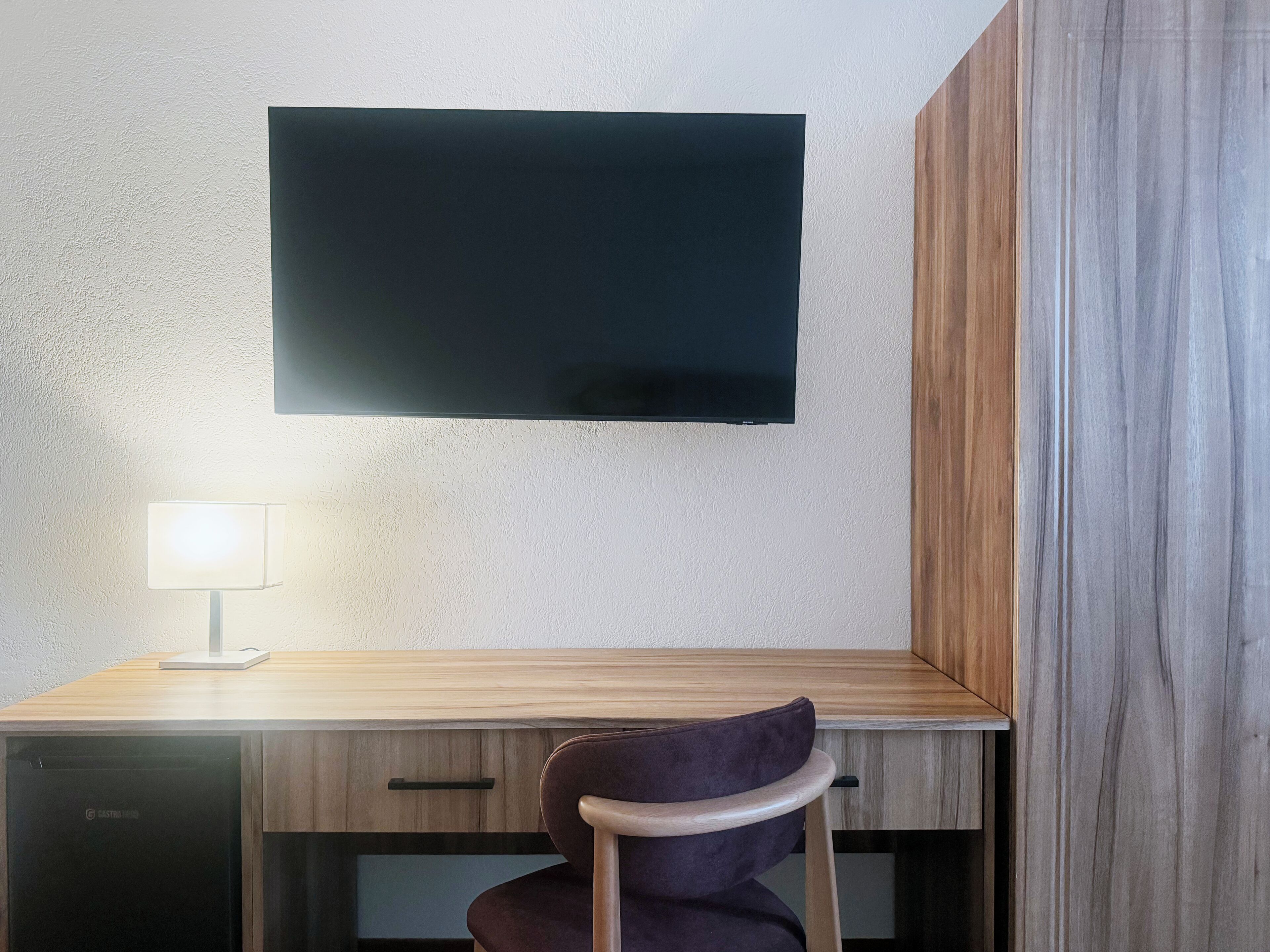 Una camera moderna con una scrivania in legno, una TV a schermo piatto fissata al muro, una lampada e una sedia.