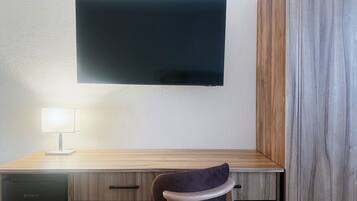 Una camera moderna con una scrivania in legno, una TV a schermo piatto fissata al muro, una lampada e una sedia.