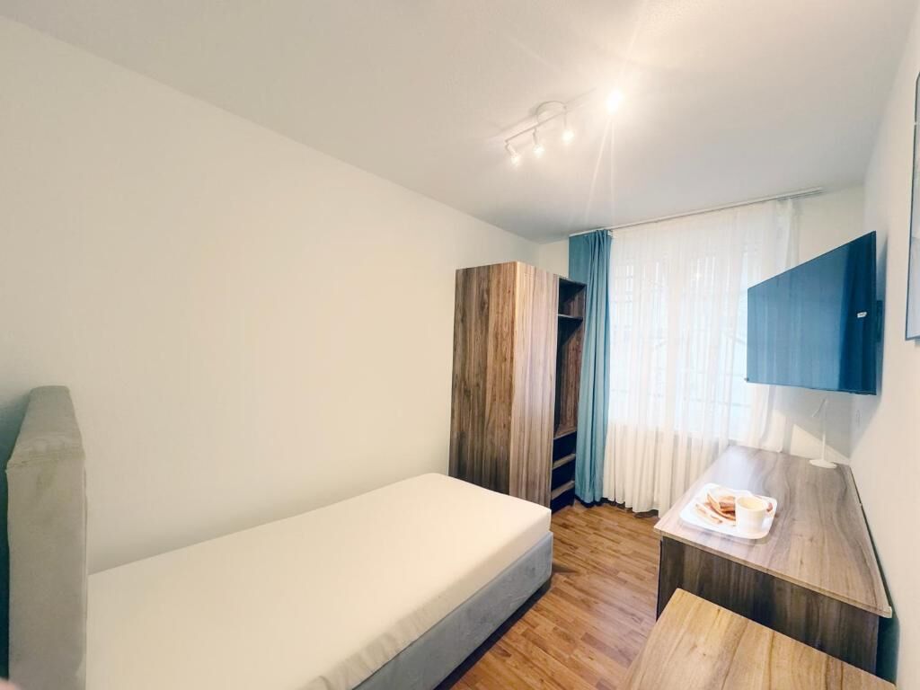 Quarto pequeno e moderno com uma cama de solteiro, escrivaninha com cadeira, televisão na parede e guarda-roupa.