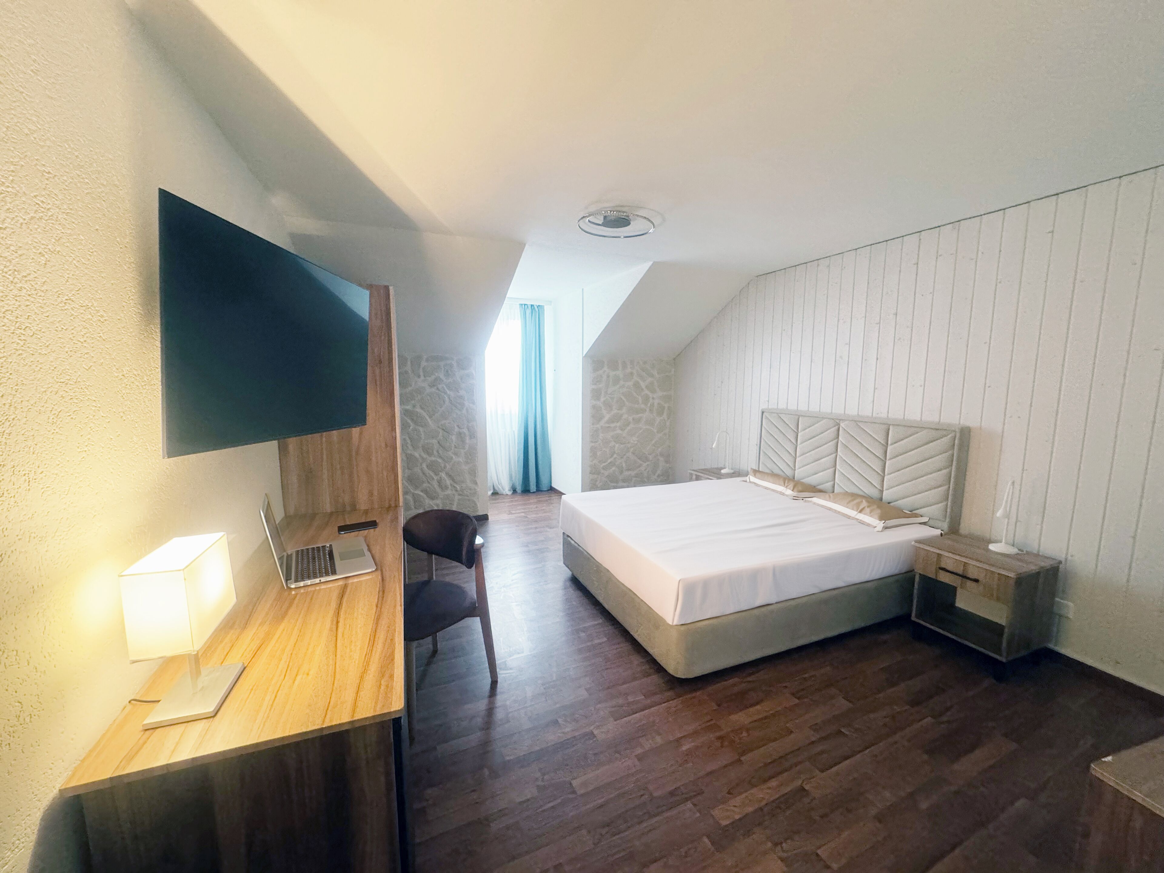 Quarto de hotel com escrivaninha de madeira, televisão de tela plana, cama com cabeceira branca e parede de pedra.