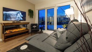 A modern living room with a large flat-screen TV, a fireplace, a grey sofa, and a balcony with a view. - OSO Summit - Downtown Riverfront Condo with Mountain Views (Golden)