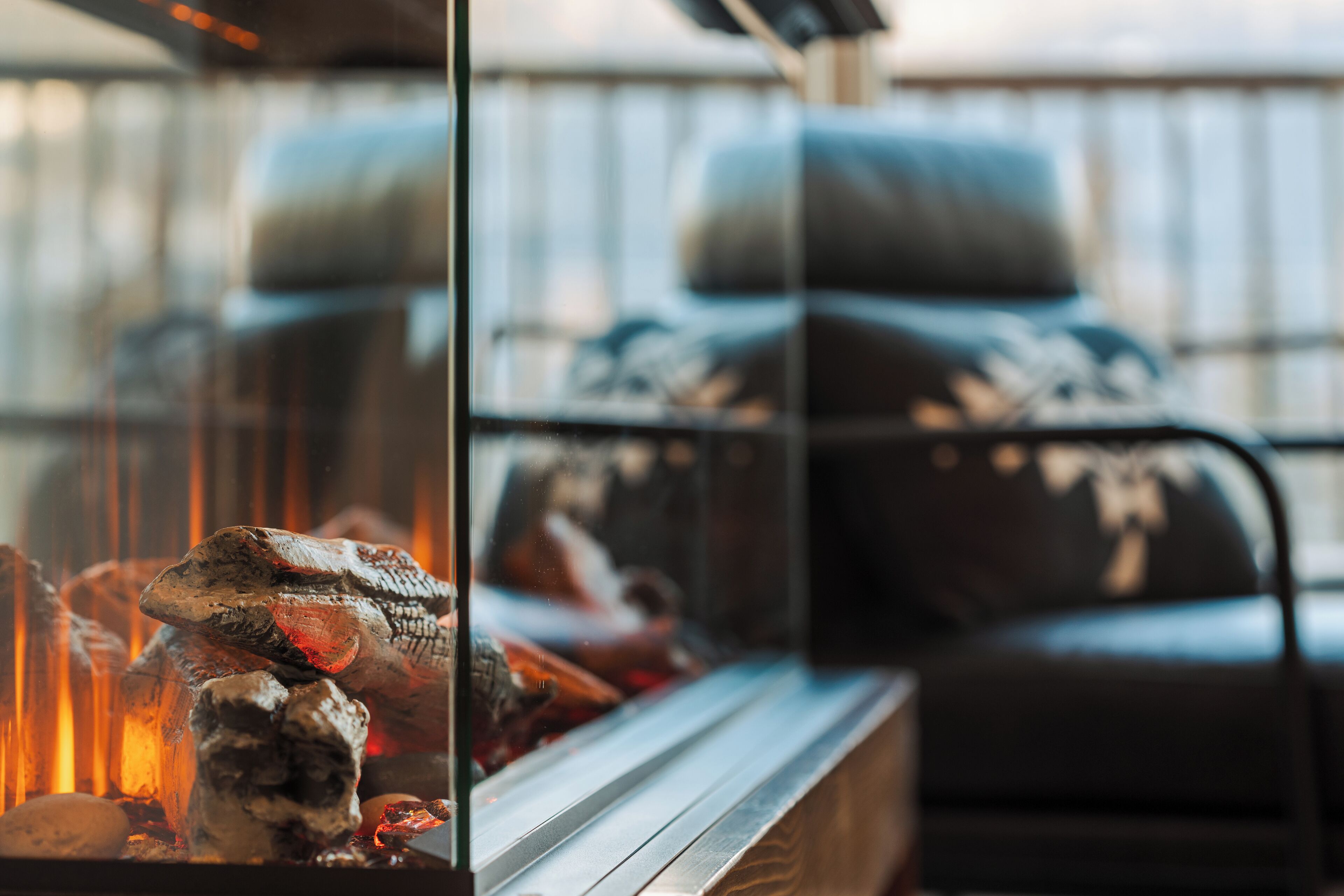 Una estructura de cristal con chimenea, una zona de estar con cojines y una barandilla de metal.