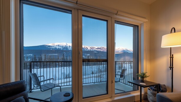 A room with large windows, a balcony with chairs, a small table, and a lamp. - OSO Summit - Downtown Riverfront Condo with Mountain Views (Golden)