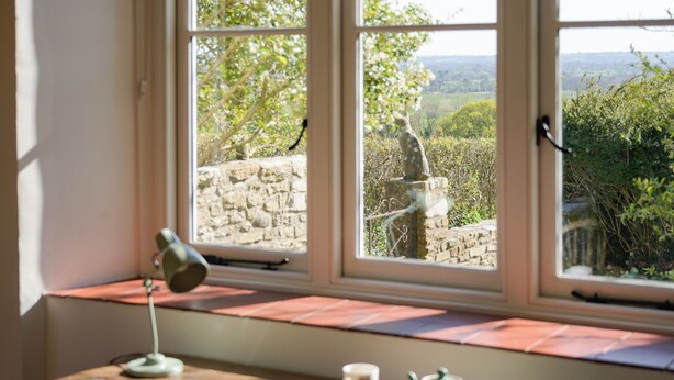 A room with a desk, a lamp, a mug, and a teapot. There is a window with a view of a stone wall and greenery.