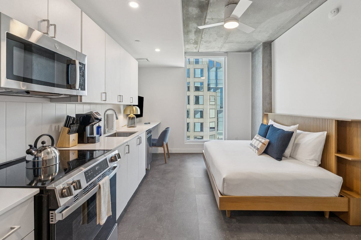 Un dormitorio pequeño con cama, kitchenette y vista a un edificio a través de una ventana.