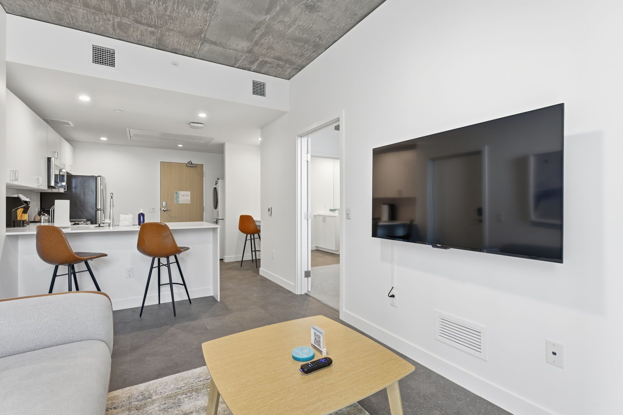 A modern living room with a flat-screen TV, a coffee table, and a sofa. There is a kitchen area with a refrigerator and a microwave in the background.