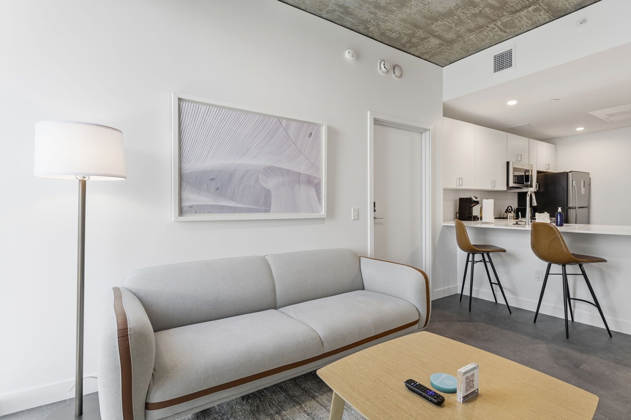 A modern living room with a sofa, a coffee table, and a lamp. There is a kitchen area with a microwave, refrigerator, and bar stools.