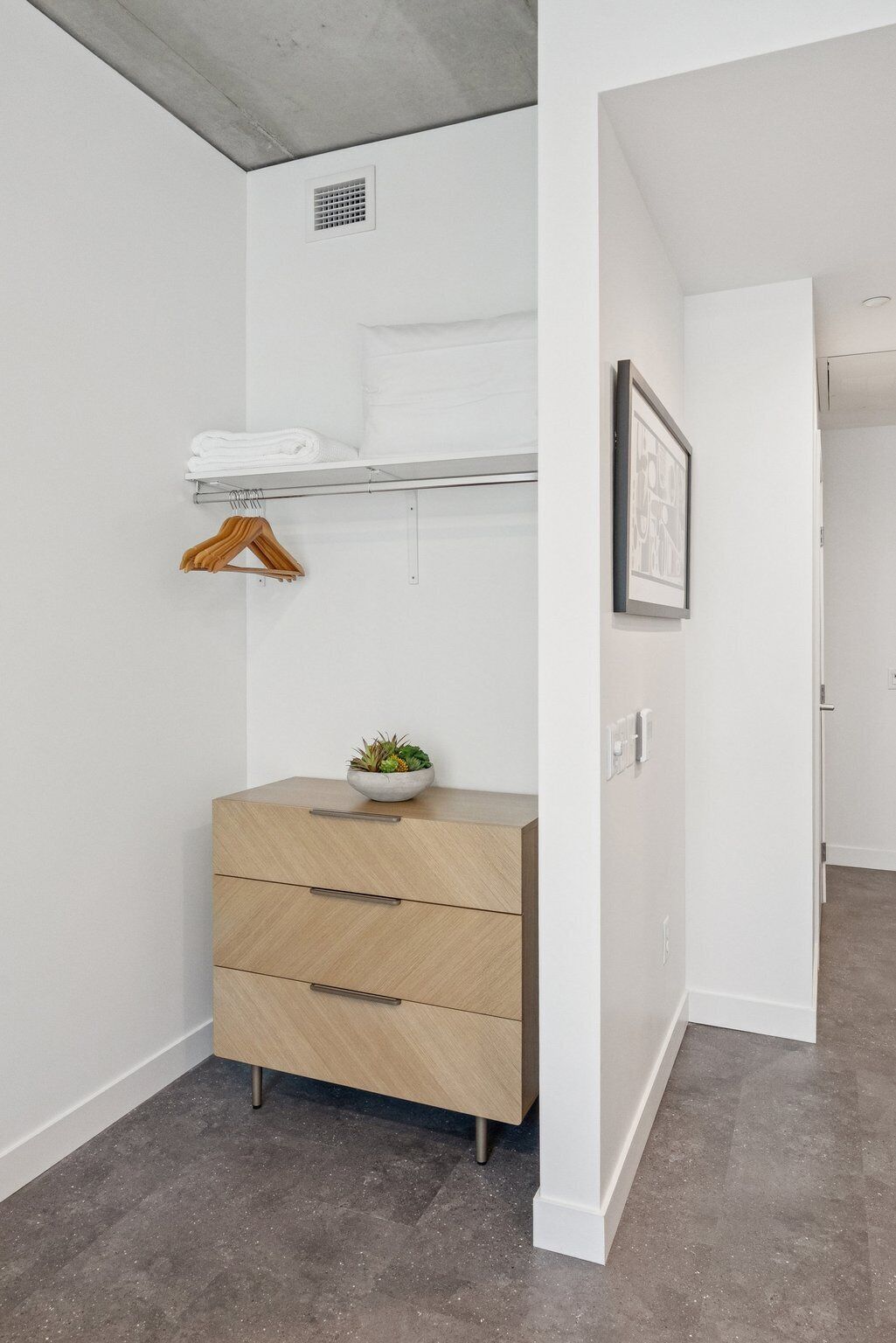 Une chambre moderne et minimaliste, avec une commode en bois, une étagère où sont pliées des serviettes, un tableau encadré et une plante en pot.