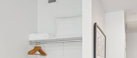 Une chambre moderne et minimaliste, avec une commode en bois, une étagère où sont pliées des serviettes, un tableau encadré et une plante en pot.