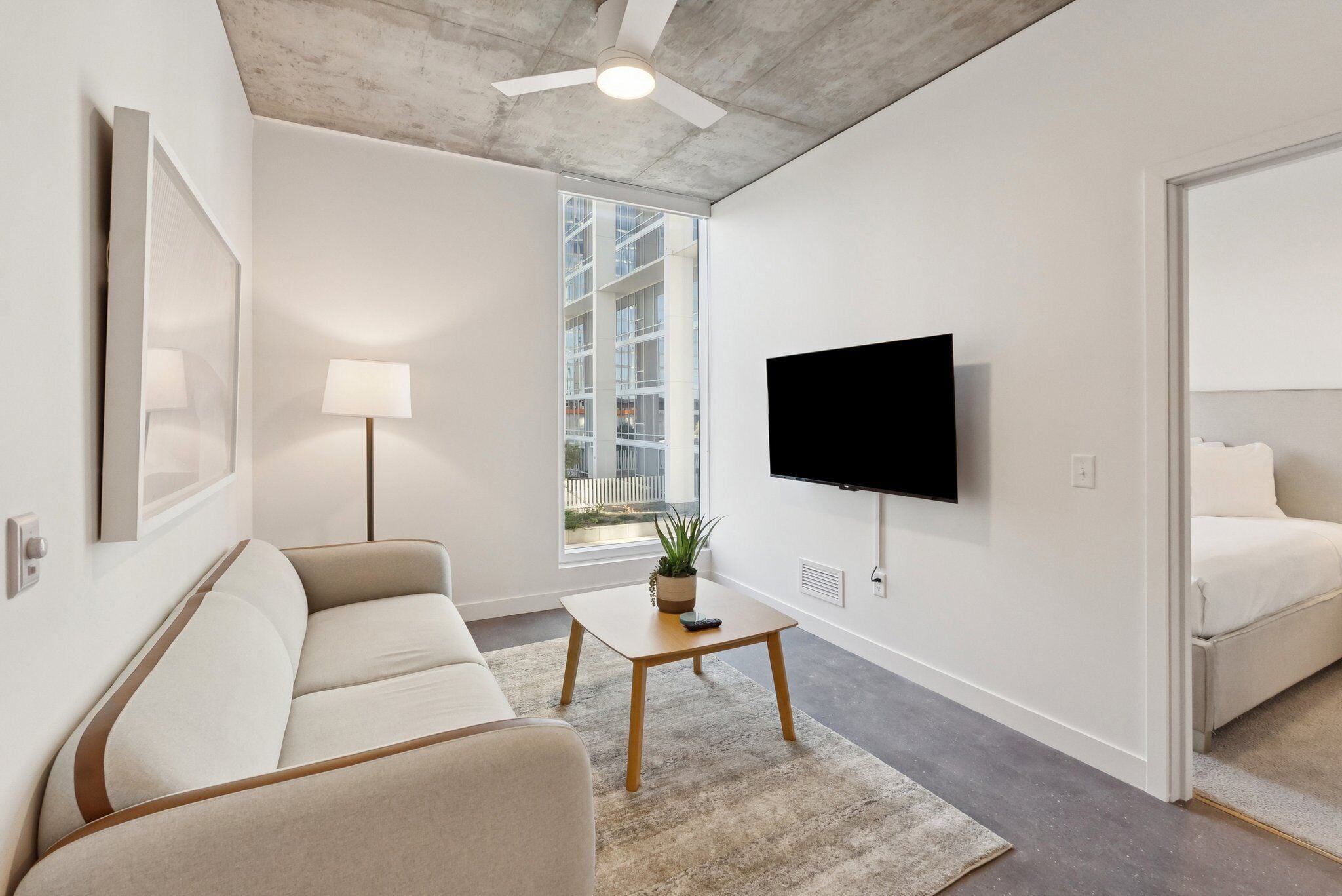Un salon moderne avec un canapé, une table basse, un téléviseur fixé au mur et une grande fenêtre donnant sur un immeuble.