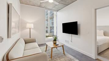 Un salon moderne avec un canapé, une table basse, un téléviseur fixé au mur et une grande fenêtre donnant sur un immeuble.
