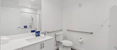 A modern bathroom with a white sink, mirror, and toilet. It includes a shower area with a glass door and handrails.