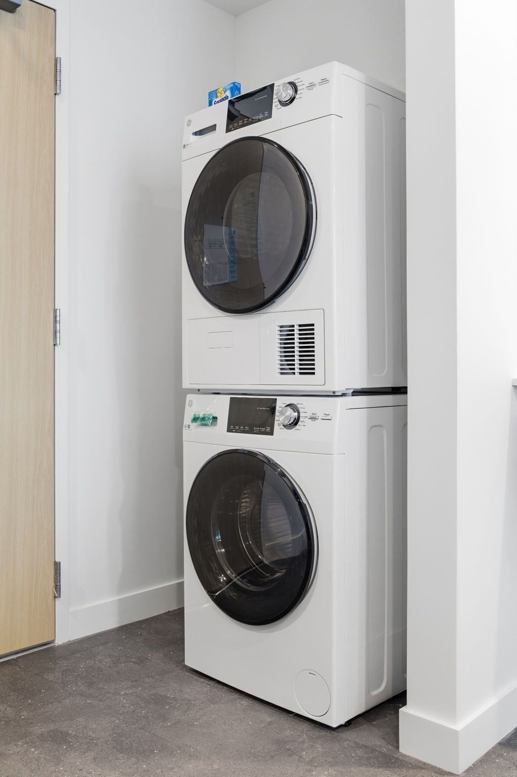 A stacked washer and dryer unit in a corner with a wooden door and a white wall.