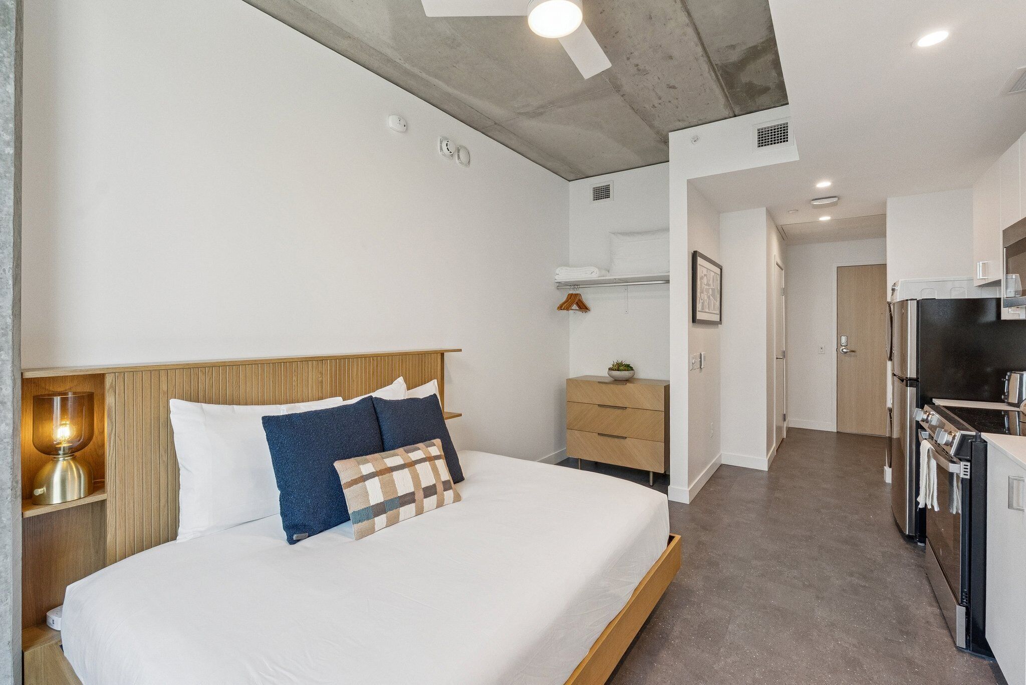 A modern hotel room with a large bed, bedside table, lamp, and a kitchenette area with a refrigerator and oven.
