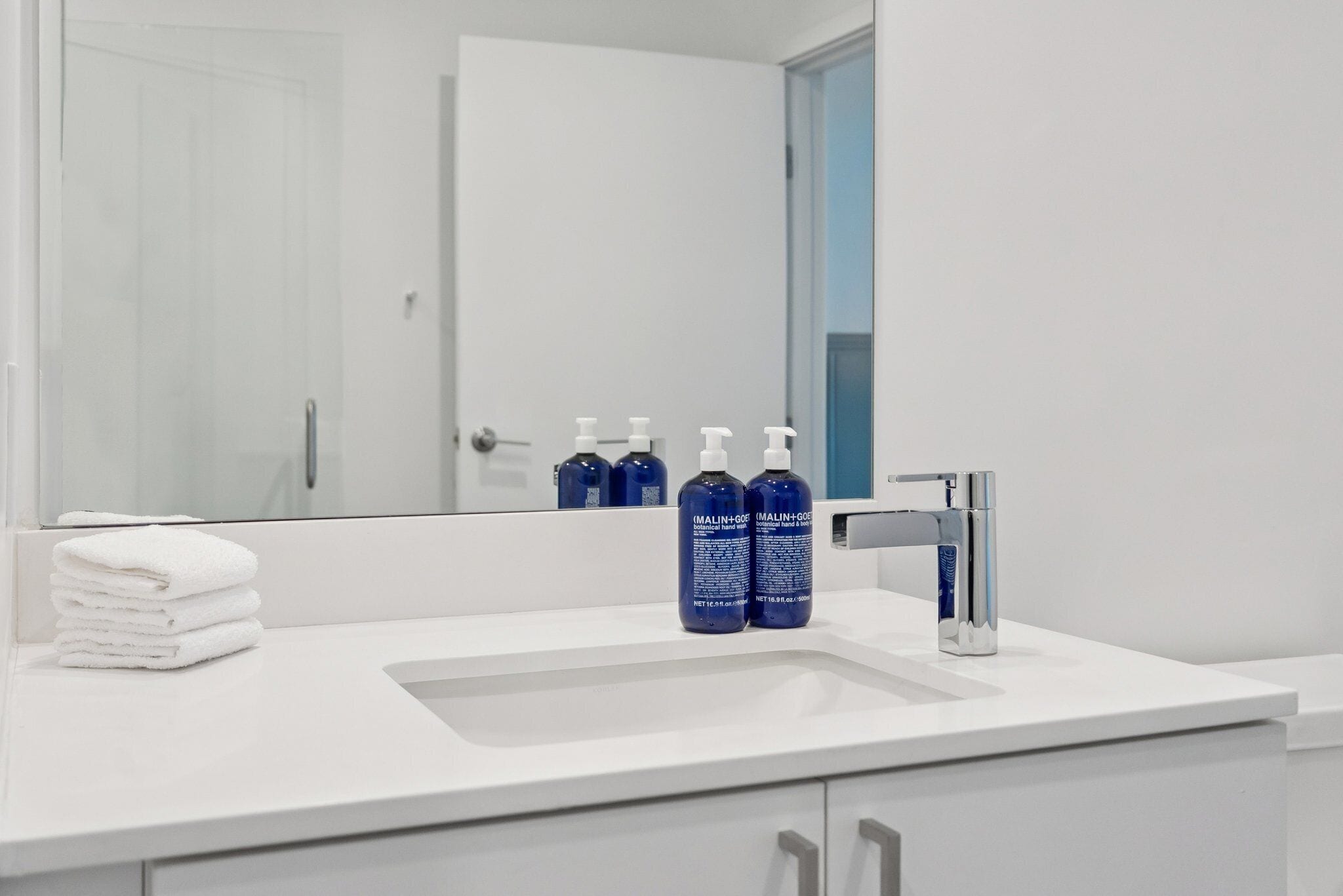 A bathroom with a white sink, two bottles of blue hand soap, a stack of white towels, and a chrome faucet.