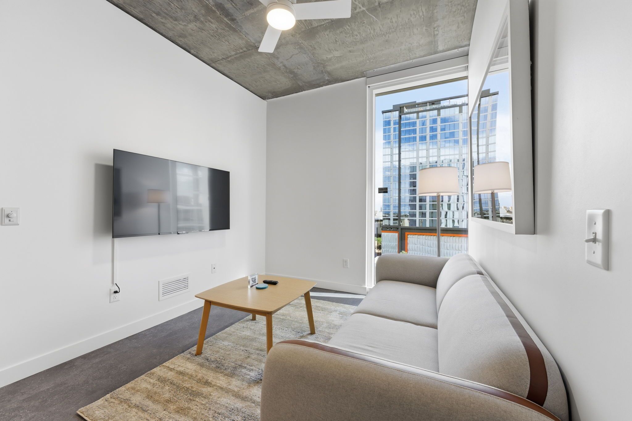 Una sala de estar moderna con un televisor de pantalla plana montado en la pared, una mesa de centro de madera, un sofá beige y un amplio ventanal con vistas a un paisaje urbano.