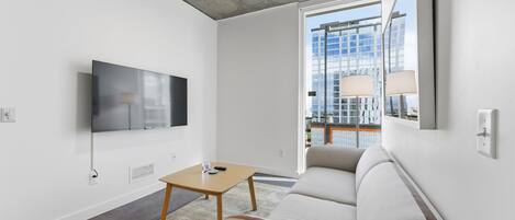 Una sala de estar moderna con un televisor de pantalla plana montado en la pared, una mesa de centro de madera, un sofá beige y un amplio ventanal con vistas a un paisaje urbano.