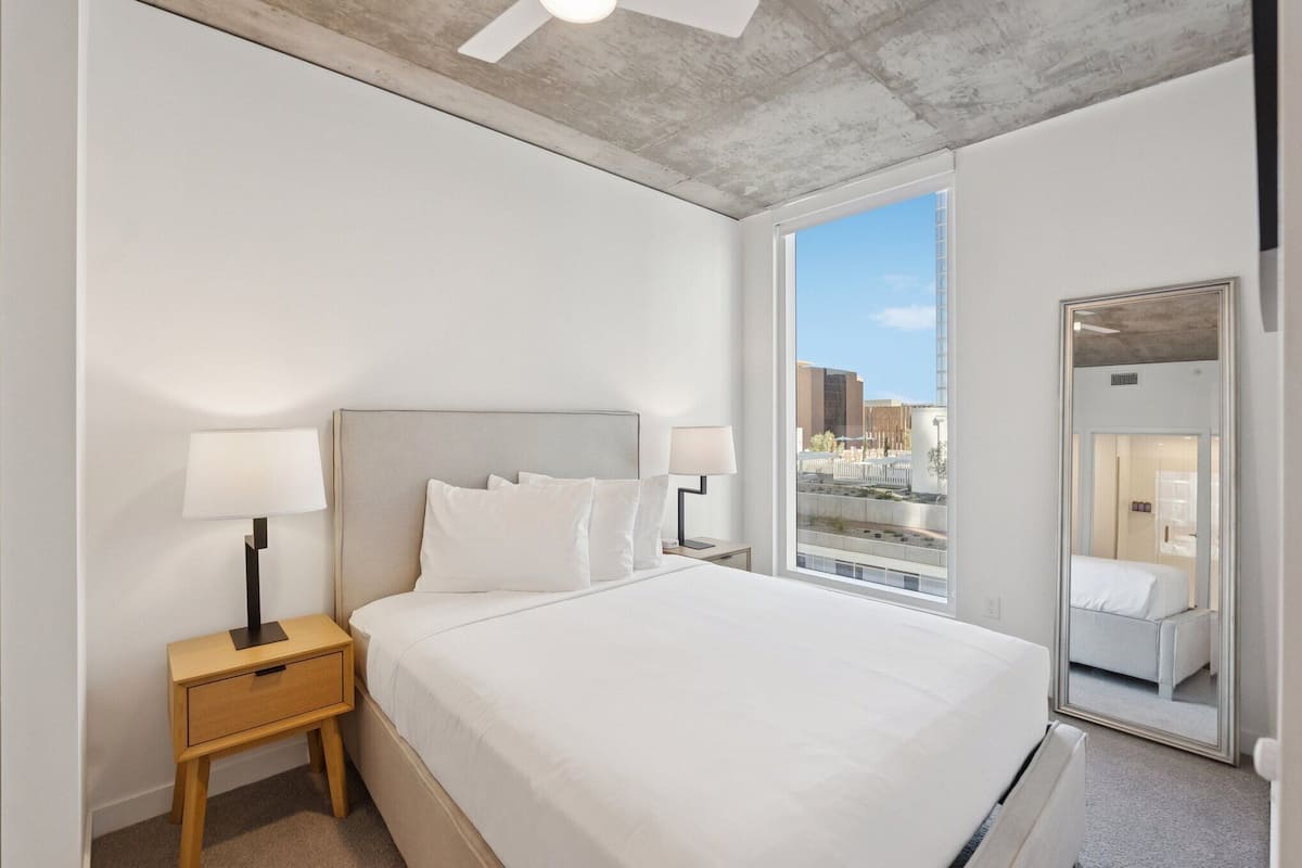 Una habitación de hotel moderna con una cama grande, dos mesitas de noche con lámparas, un ventanal con vistas a la ciudad y un espejo en la pared.