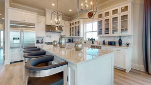 A spacious kitchen with white cabinetry, a central island with bar stools, stainless steel appliances, and decorative items. - 2nd Row Stunner w/ Gulf Views, Steps to the Beach! (Galveston)