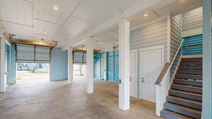 A spacious indoor area with concrete floors, white pillars, and a staircase with wooden steps and blue shutters. - 2nd Row Stunner w/ Gulf Views, Steps to the Beach! (Galveston)