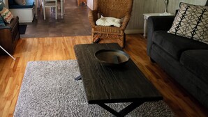Ein Wohnzimmer mit einem dunklen Holztisch, einem grauen Sofa, einem Korbstuhl und einem Essbereich im Hintergrund.