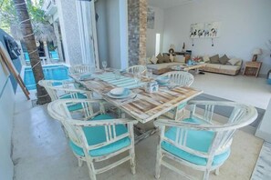 A dining area with a wooden table and white chairs, a pool, and a living area with a sofa and cushions.