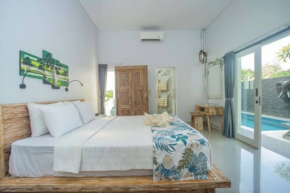 Un dormitorio con cama, cabecera de madera, ventana con vista a una piscina y puerta que da a otra habitación.