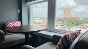 Una habitación con una mesa redonda, dos sofás grises y una ventana con vistas a edificios y árboles.