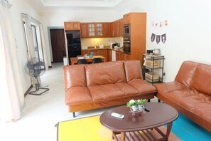 A living room with a sofa, a coffee table, and a fan. There is a kitchen in the background with wooden cabinets and appliances.