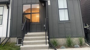 A two-story house with a dark gray exterior, a front entrance with a glass door, and a small front yard with grass and plants. - Cozy quiet place 15 min driver to airport perfect place for family and friends. (Calgary)