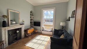 Una sala de estar con chimenea, un sofá, una mesa de centro, una estantería y un amplio ventanal con vistas a edificios.