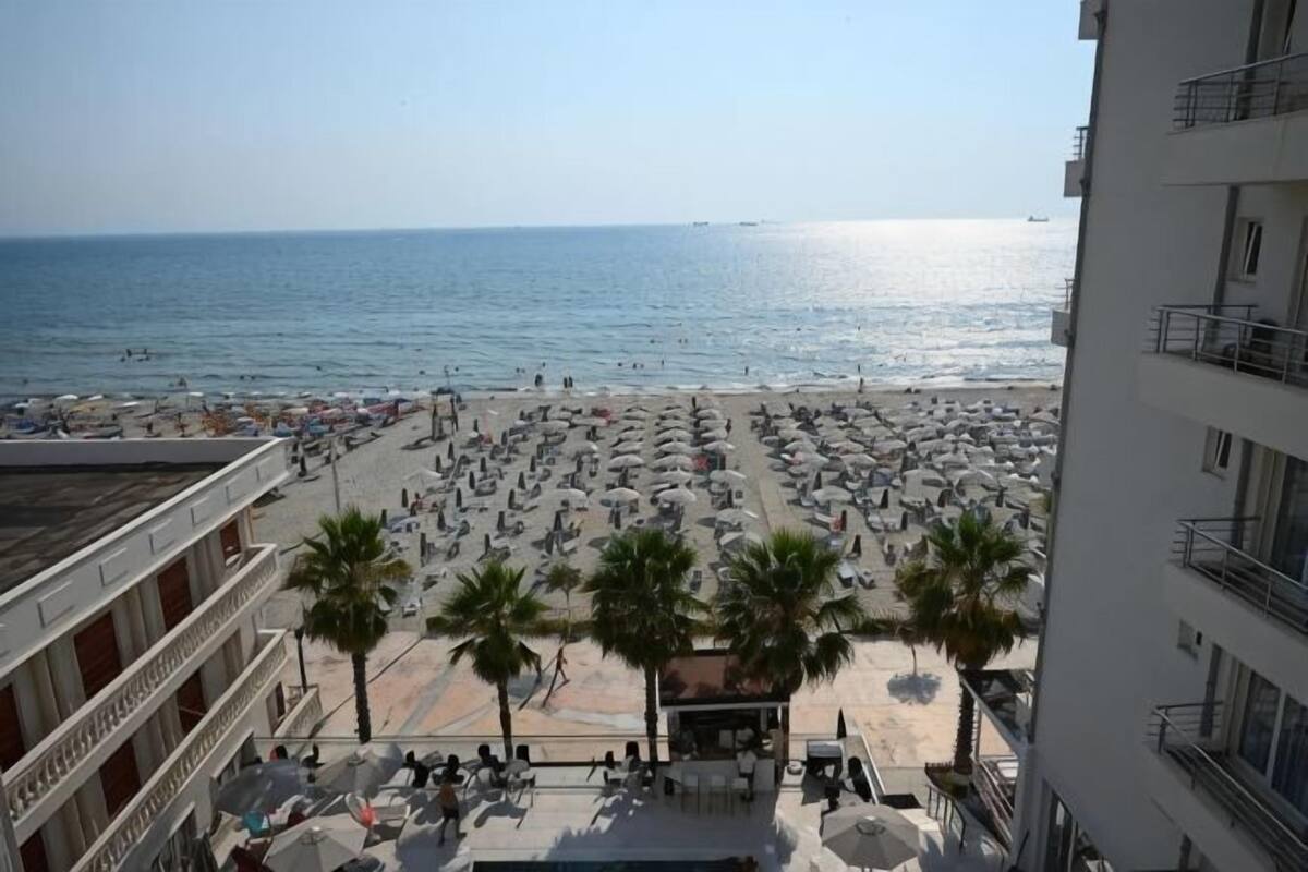 Une vue sur la plage depuis le balcon d’un hôtel à plusieurs étages, avec des palmiers et un ciel dégagé.